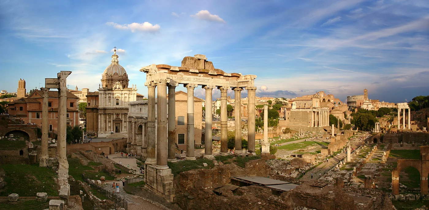 Panoramic view of the Roman Forum archaeological site showing ancient temple ruins, columns, and the Via Sacra with the Colosseum visible in the background