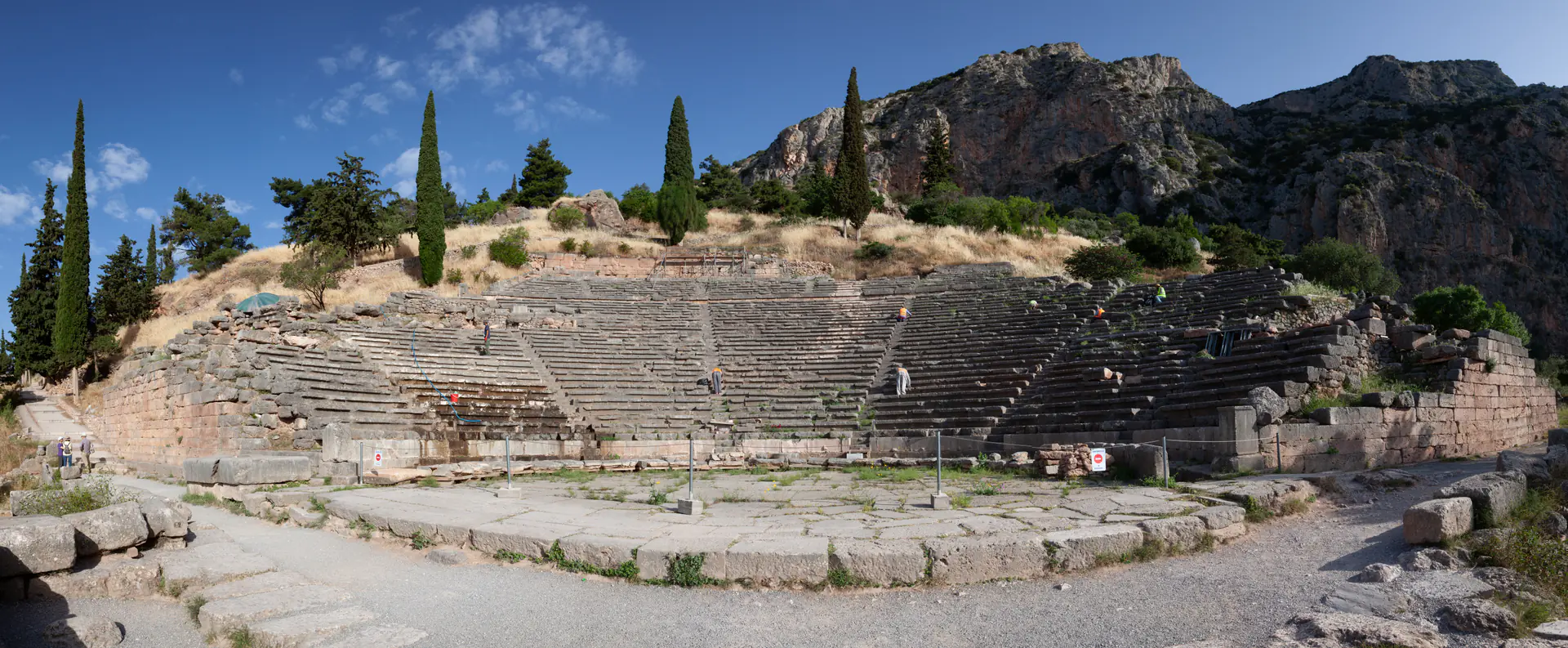 Ultra-wide panoramic view of the ancient theatre at Delphi showing the curved stone seating tiers carved into Mount Parnassus with the dramatic valley of Phocis and mountain ranges stretching to the horizon