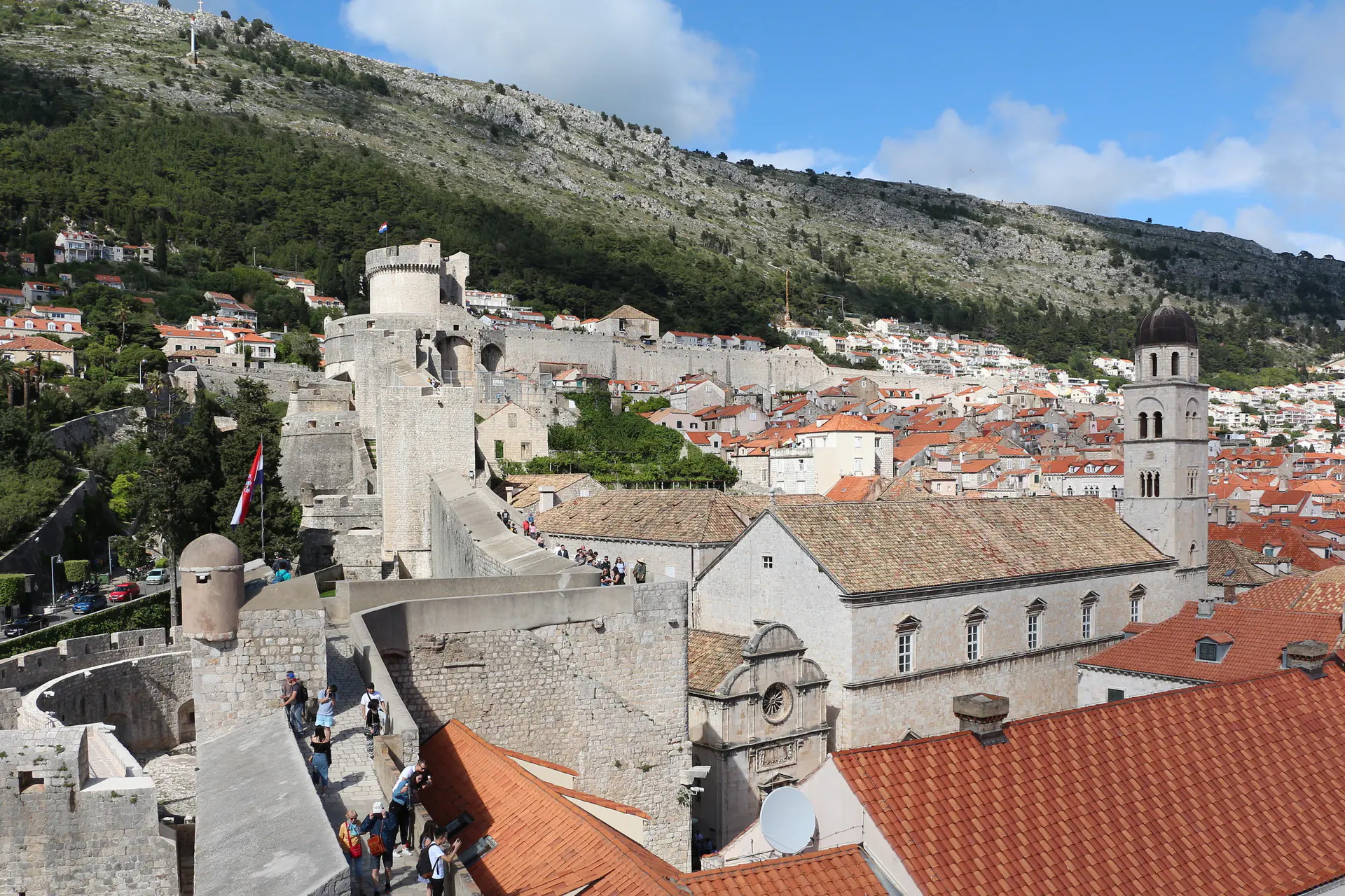 Vista panorámica a lo largo de las murallas medievales de piedra de Dubrovnik mostrando torres de vigilancia, pasarela con turistas, y tejados de terracota abajo con el mar Adriático más allá