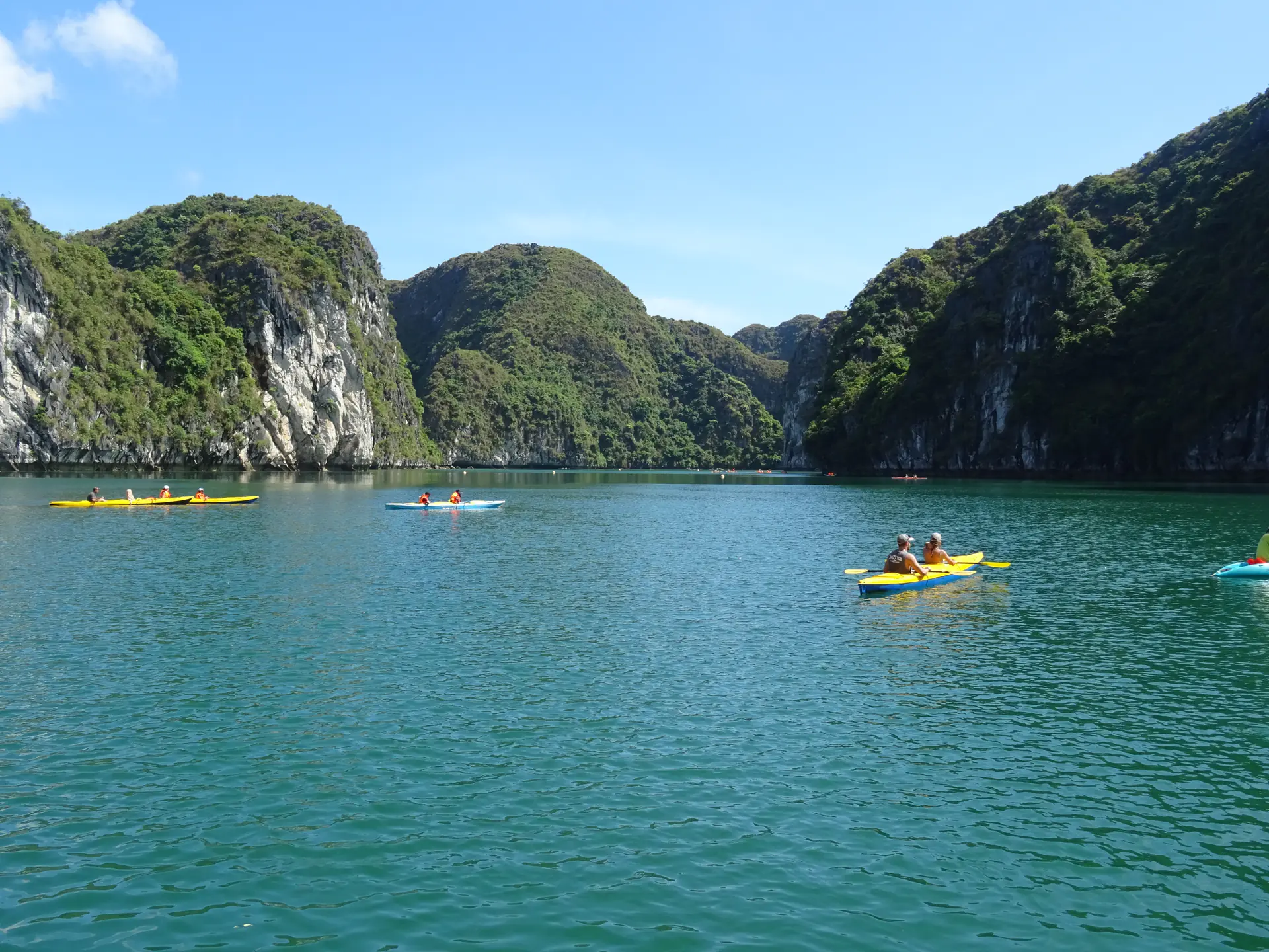 Kayakistas remando a través de aguas esmeralda tranquilas entre imponentes formaciones kársticas de piedra caliza en la Bahía de Ha Long con vegetación exuberante aferrándose a las caras de los acantilados