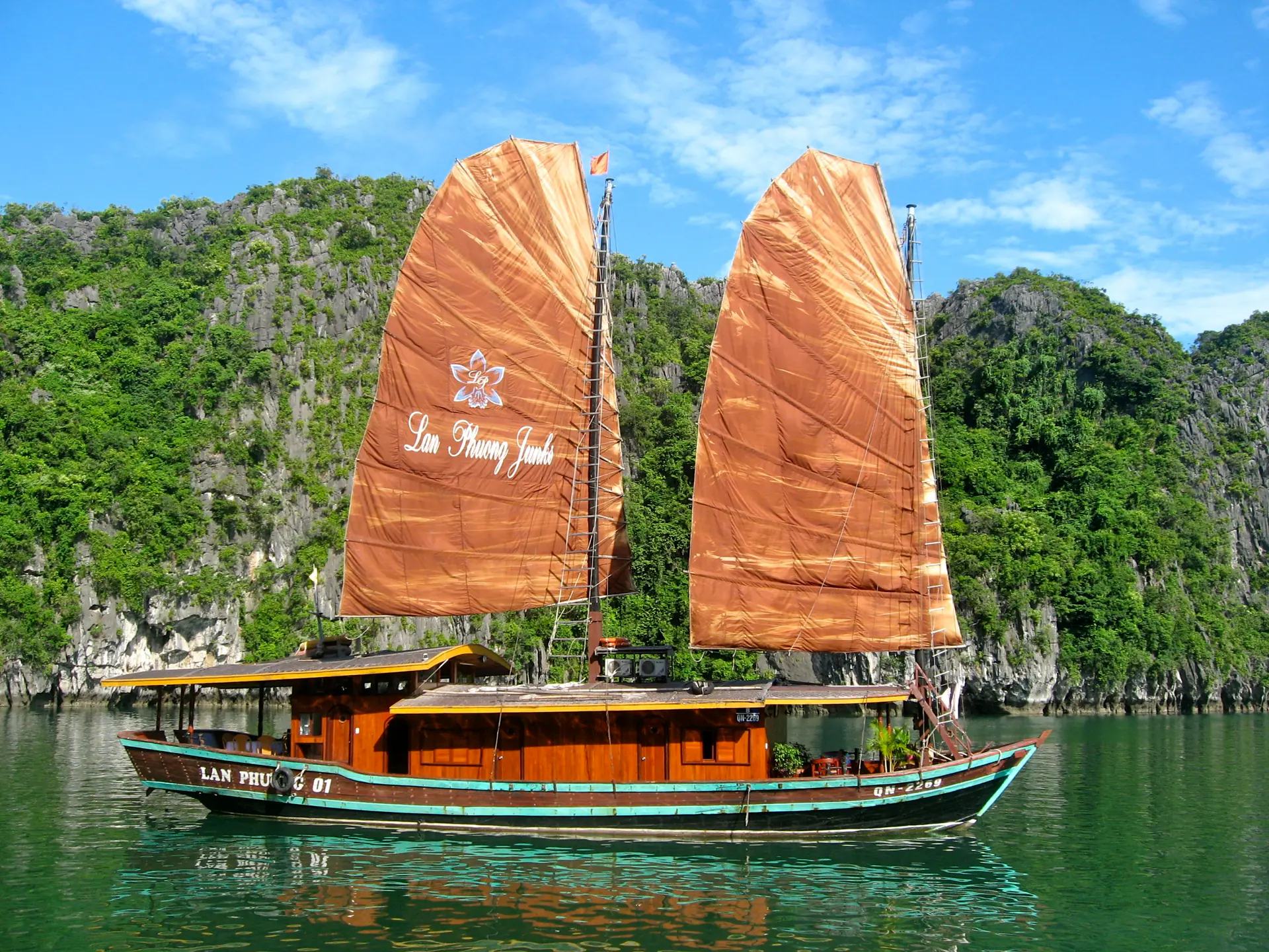 Barco junk vietnamita tradicional con velas rojas navegando por la Bahía de Ha Long con dramáticas formaciones kársticas de piedra caliza elevándose desde aguas esmeralda al fondo