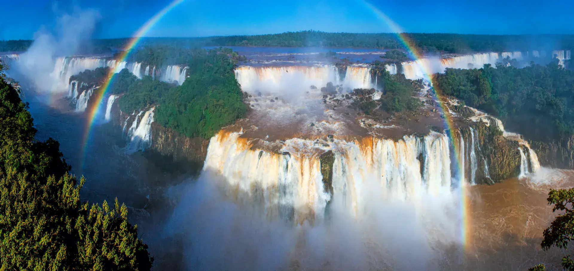 Spectacular aerial view of Iguazu Falls with rainbow formed by waterfall spray, showing the complete falls system from above with lush Atlantic Forest surrounding the cascades