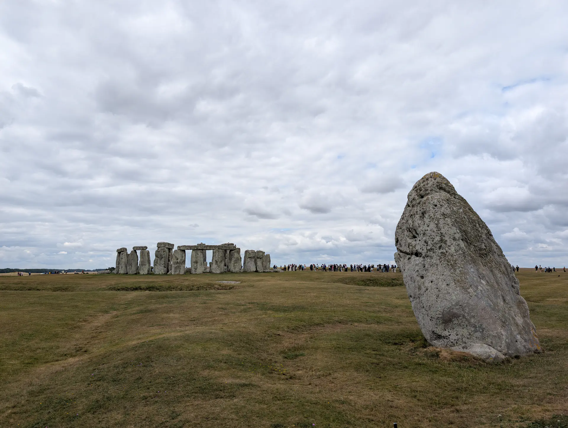 The Heel Stone standing prominently in the foreground with Stonehenge stone circle visible in the background across the grass field