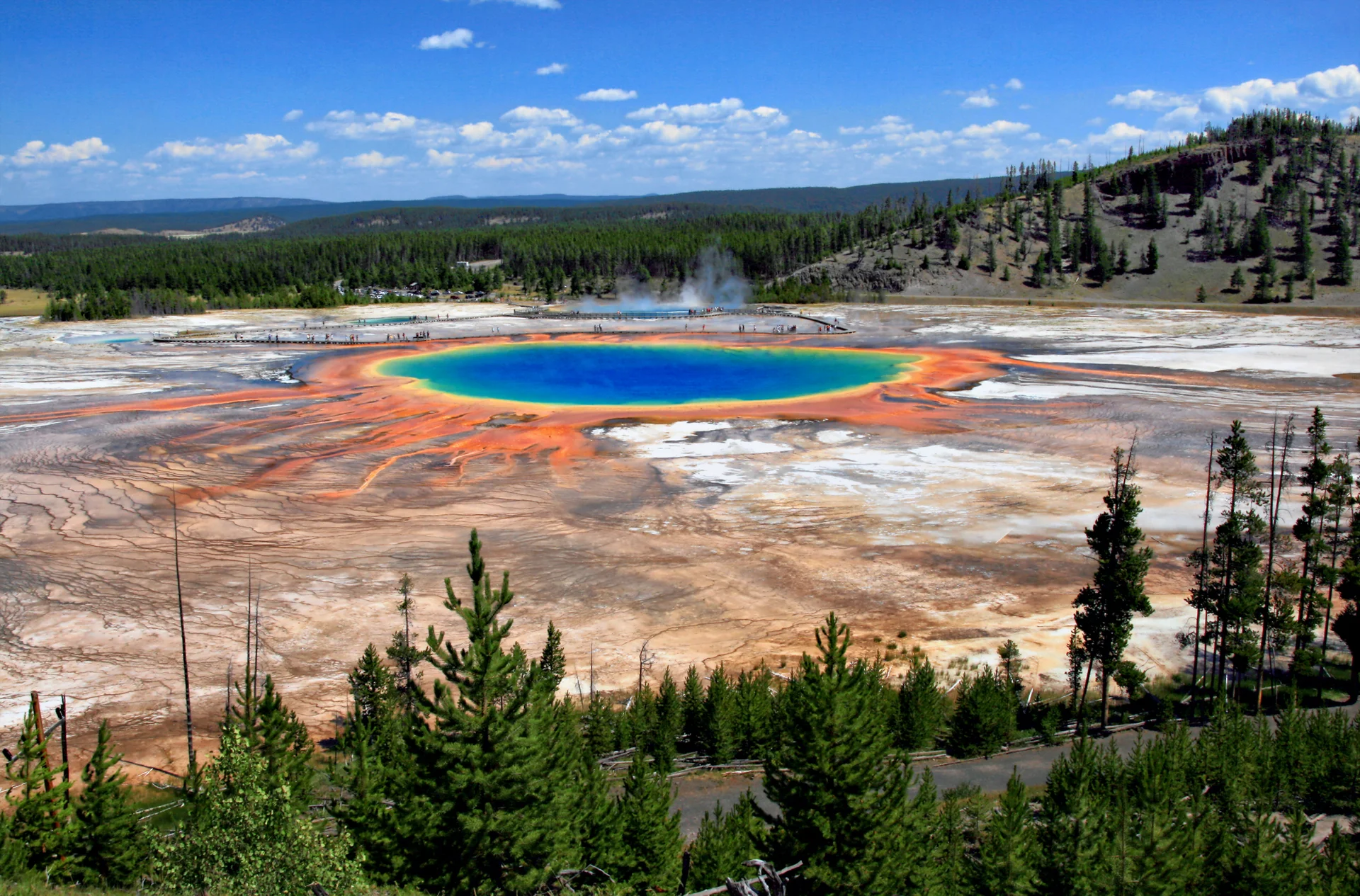 Aerial view of Yellowstone's Grand Prismatic Spring showing vibrant rainbow colours radiating from the deep blue centre through orange and yellow bacterial mats